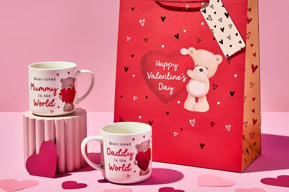 Red Valentine’s Day gift bag with a teddy bear design and heart patterns, accompanied by two white mugs with red text reading Most-loved Mummy in the World and Most-loved Daddy in the World, surrounded by scattered paper hearts on a pink background.